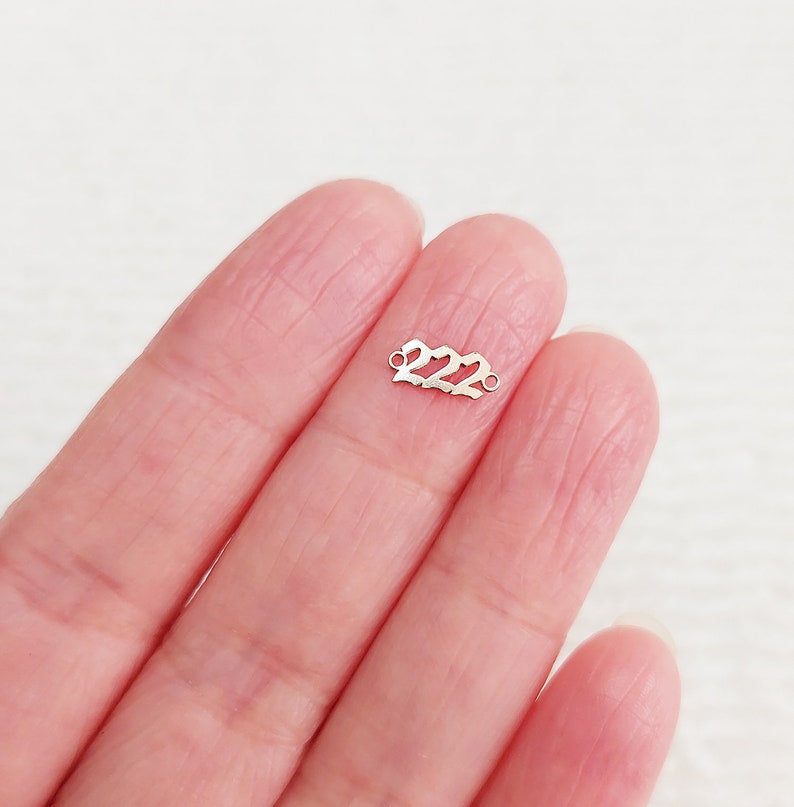 Może przedstawiać: Mały, srebrny charms z wyciętą liczbą "222". Charms ma dwie małe pętle na każdym końcu do mocowania do biżuterii. Charms jest trzymany w dłoni osoby.