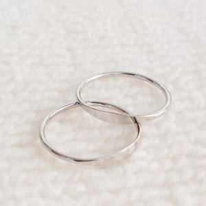 May include: Two silver rings, one overlapping the other, against a white, textured background. The rings are thin and have a polished, reflective surface. The rings appear to be made of metal.