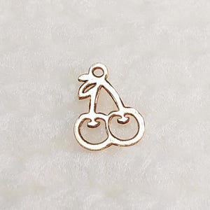 May include: A gold-colored cherry charm with a loop at the top for attaching to jewelry. The charm features a detailed outline of two cherries connected by a stem and leaves. The charm is set against a white background.