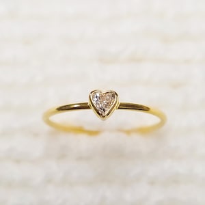 May include: A delicate gold ring featuring a heart-shaped diamond. The ring's band is thin and polished, leading to the bezel-set diamond. The diamond has a clear, sparkling appearance. The ring is a symbol of love and commitment.
