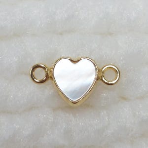 May include: A gold-colored heart-shaped connector with a pearlescent center. The connector has two small loops on either side. The heart shape is outlined in gold.