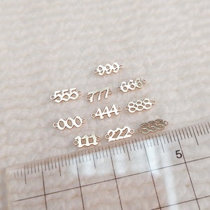 Może przedstawiać: Kolekcja małych, złotych metalowych zawieszek, każda z innym wzorem liczby. Numery obejmują 000, 111, 222, 444, 555, 666, 777, 888 i 999. Na zdjęciu znajduje się linijka.