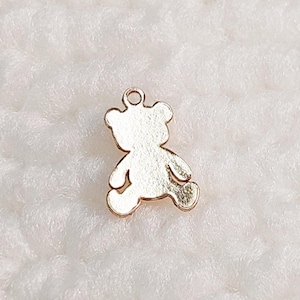 May include: A gold-colored teddy bear charm with a loop at the top for attaching to jewelry. The bear is in a playful pose, with its arms and legs slightly outstretched. The charm is made of a shiny metal and has a smooth surface.