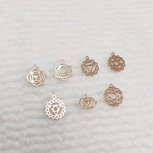 Peut inclure: Sept breloques dorées représentant les chakras, avec des motifs géométriques complexes. Chaque breloque présente un motif ajouré unique, comprenant des cercles, des triangles et des formes florales. Les breloques sont petites, avec une boucle pour la fixation.