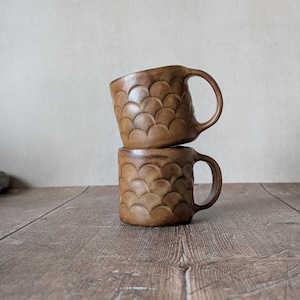 May include: Two stacked brown ceramic mugs with a textured, scalloped design. The mugs have a handle and a warm, earthy tone, set against a neutral background on a wooden surface. The mugs are approximately 10 cm tall.