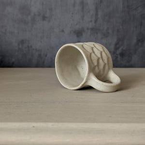 May include: A speckled off-white ceramic mug resting on its side. The exterior surface features a raised, repeating scale-like pattern. The mug has a curved handle and a smooth, rounded rim, showcasing a natural, earthy clay texture throughout.