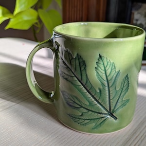 May include: A light green ceramic mug featuring a prominent embossed leaf design on the exterior. The mug has a smooth, glossy finish with a curved handle on the side. The leaf pattern displays detailed veins in a darker shade of green.
