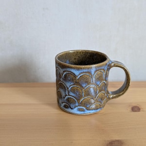 May include: A ceramic mug with a brown and blue glaze. The mug has a textured, scale-like pattern on the exterior. The handle is brown, and the interior has a light blue colour. The mug is on a wooden surface.