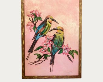 May include: A framed painting featuring two colourful birds with long, curved beaks perched on branches with pink flowers. The birds have blue, green, and yellow plumage. The background is a textured, soft pink, and the frame is a metallic gold finish.