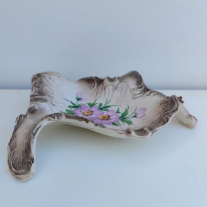 Pode incluir: Um prato de cerâmica branca com uma borda festonada e um design floral. O prato tem um esmalte marrom e tem a forma de uma folha. As flores são rosa e amarelas.