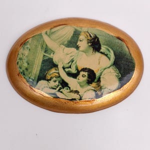 Pode incluir: Moldura de madeira oval com acabamento dourado, apresentando uma impressão vintage de uma mulher e duas crianças. A mulher está vestindo um vestido branco e amarelo, e as crianças estão vestindo vestidos brancos. A impressão é sobre um fundo verde.