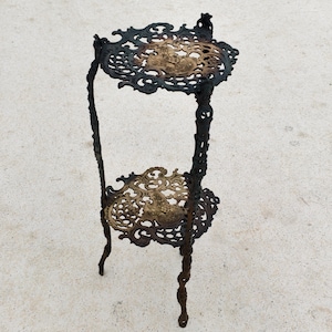 Könnte beinhalten: Verzierter, zweistöckiger Pflanzenständer mit dunkelbronzenem Finish. Der Ständer weist auf den kreisförmigen Ablagen und Verbindungsstützen komplizierte, durchbrochene Designs auf. Die obere Ablage hat einen Durchmesser von ca. 25 cm, die untere Ablage ca. 30 cm.