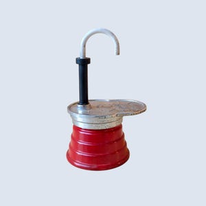 Puede incluir: Una cafetera de metal rojo de estilo vintage con base escalonada, sección central metálica plateada y un vástago vertical negro rematado con un asa blanca curva. La tapa plateada presenta un logotipo circular en relieve con texto.