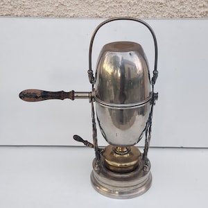 May include: A silver and brass coffee percolator with a wooden handle. The percolator has a round, bulbous shape and sits on a stand with a decorative base.