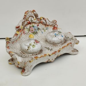 Puede incluir: Un tintero de cerámica blanca con un diseño floral decorativo y dos pequeños recipientes con tapa. El tintero tiene un esmalte craquelado y un aspecto envejecido.