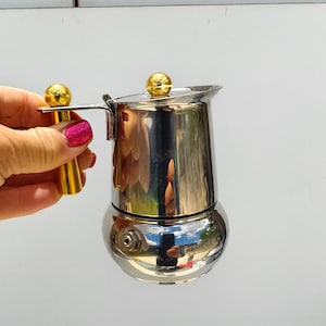 Peut inclure: Une petite cafetière argentée brillante avec une poignée et un bouton supérieur dorés. La cafetière a une base ronde et un corps cylindrique. La poignée et le bouton supérieur sont dorés. La cafetière est tenue par une personne.