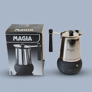 May include: A stainless steel Magia stovetop espresso maker with a black base and handle, accompanied by its original black cardboard box. The box features the Magia logo and a graphic of the coffee maker. The unit is labelled 6 cup 18/10 stainless steel.