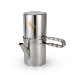 Könnte beinhalten: Eine zylindrische Kaffeemaschine aus Edelstahl mit polierter Oberfläche, ausgestattet mit einem seitlichen Griff und einem kleinen Ausguss. Ein rechteckiges Etikett mit der Aufschrift STELLA befindet sich am oberen Teil. Der Deckel ist abnehmbar.