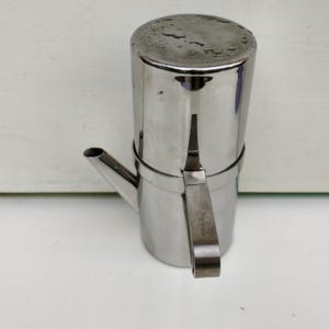 May include: A stainless steel stovetop espresso maker with a cylindrical body, a spout, and a handle. The coffee pot has a shiny, reflective surface and is designed for brewing espresso. The handle is attached with a metal band.