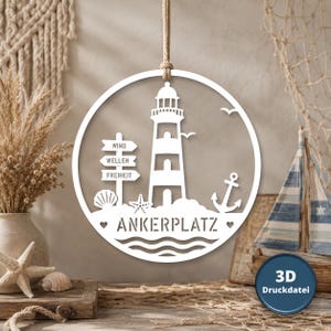 Könnte beinhalten: Eine weiße, kreisförmige Wanddekoration zum Aufhängen mit einem Leuchtturm, einem Wegweiser mit den Wörtern WIND, WELLEN, FREIHEIT, einer Muschel, einem Seestern, einem Anker und dem Schriftzug ANKERPLATZ, aufgehängt an einem dicken Seil.