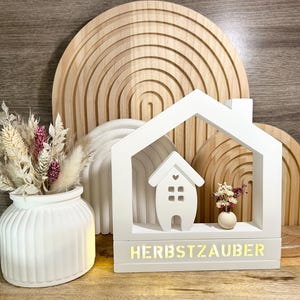 Könnte beinhalten: Weißes, hausförmiges Licht aus Holz mit einem kleinen Haus-Ausschnitt im Inneren und dem Text "HERBSTZAUBER" auf der Basis. Das Licht steht auf einer Holzoberfläche vor einem Holzregenbogen.