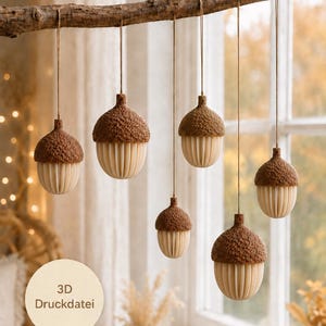 May include: Six hanging acorn ornaments suspended from a horizontal wooden branch. Each acorn features a textured brown cap and a cream-colored, vertically ribbed base. A circular label in the lower left corner displays the text 3D Druckdatei.