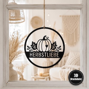 May include: A circular black hanging wall decoration featuring a central pumpkin graphic with leaves and a small heart cutout. The word HERBSTLIEBE is printed in a bold, sans-serif font across a horizontal bar. A small black circle with 3D Druckdatei text.