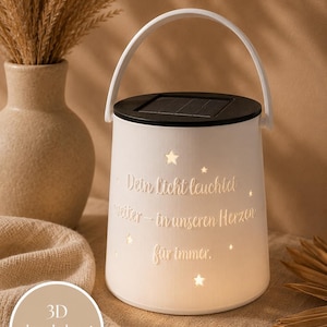 May include: A white cylindrical solar lantern with a handle and a black top panel. The lantern features cut-out star shapes and the German text "Dein Licht leuchtet weiter - in unseren Herzen für immer." The surface emits a soft internal glow.