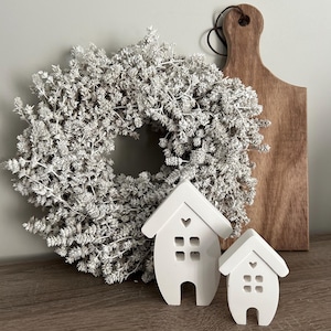 Könnte beinhalten: Zwei weiße Keramikhäuser mit Herzdetails und Fensterschnitten. Die Häuser stehen auf einer Holzoberfläche vor einem weißen Kranz.