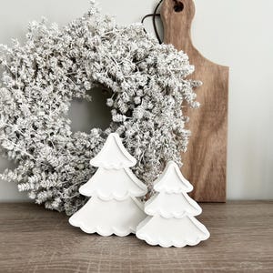 Può includere: Due decorazioni per albero di Natale in legno bianco con un design a bordo smerlato. Gli alberi sono su una superficie di legno.