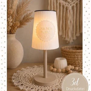 Puede incluir: Una lámpara de mesa con base beige y pantalla cónica blanca con rayas verticales. La pantalla presenta un emblema circular con borde festoneado que contiene el texto "LIEBLINGS PLATZ" y un pequeño símbolo de corazón. La lámpara emite luz.