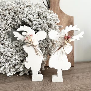 Peut inclure: Deux figurines de rennes en bois blanc avec des fleurs séchées et des accents de ficelle. Les rennes se font face et semblent s'embrasser.
