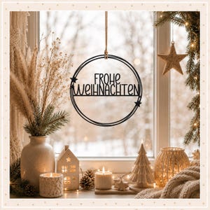 Op de afbeelding: Een zwarte, ronde hangdecoratie met de tekst "FROHE WEIHNACHTEN" in een gestileerd lettertype, versierd met twee kleine sterren aan de zijkanten. Het ornament hangt aan een dun koord en is ontworpen om voor een raam te worden opgehangen.