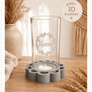 May include: A clear glass cylinder candle holder featuring a floral wreath design with the word Home etched on the front. The glass sits inside a matte gray, flower-shaped base that holds a small white tea light candle in the center.