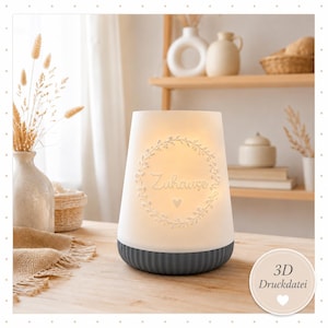 May include: A white ceramic candle holder with a gray base, featuring the word "Zuhause" within a floral wreath. The candle holder is illuminated, creating a warm glow. The design includes a heart symbol.