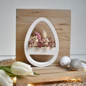 Könnte beinhalten: Weiße, eiförmige Wanddekoration mit getrockneten Blumen in Rosa- und Beigetönen. Auf einem Hintergrund aus Holzbrettern montiert. Weiße Tulpen und gesprenkelte Ostereier sind im Vordergrund.