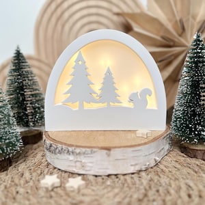 Könnte beinhalten: Eine weiße, bogenförmige Weihnachtsdekoration aus Holz mit einer Winterszene mit zwei Bäumen und einer Eichhörnchen-Silhouette, beleuchtet von warmen Lichtern. Die Dekoration steht auf einem Birkenholzsockel mit kleinen sternförmigen Akzenten.