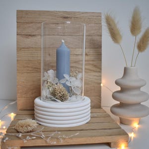 Peut inclure: Présentation décorative d'une bougie bleue pâle sous cloche en verre. La bougie repose sur une base blanche et côtelée, entourée de fleurs séchées. Un vase beige avec des plantes séchées et un fond en bois complètent la scène.