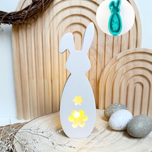 Könnte beinhalten: Weißer, hölzerner Osterhase in Form eines Eies mit gelben Blumenausschnitten. Der Hase steht auf einer Holzscheibe. Im Hintergrund befindet sich eine grüne Silikonform des Hasen.