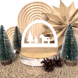 Può includere: Decorazione natalizia in legno bianco con un cavallo e alberi all'interno di un arco, su una base in legno di betulla. Alberi di Natale verdi in miniatura, un ornamento di carta a forma di stella e pigne circondano la scena.