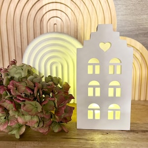 Może przedstawiać: Biała ceramiczna lampka nocna w kształcie domu z wyciętym sercem i oknami. Światło jest włączone i emituje ciepłe światło.