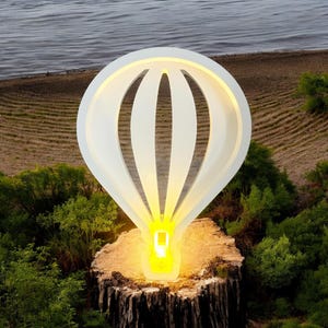 Op de afbeelding: Een witte houten sculptuur van een heteluchtballon met een gloeiend geel licht erin, die op een boomstronk in een grasachtig gebied staat.