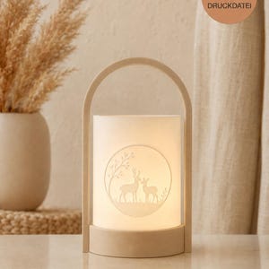 May include: A cylindrical lantern with a light beige frame and handle, featuring a translucent white shade. The shade displays a circular embossed graphic of two deer standing in a forest setting. A circular badge reads 3D DRUCKDATEI.