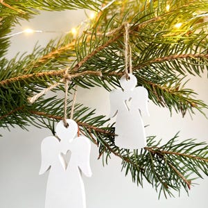 Könnte beinhalten: Zwei weiße Keramik-Engel-Anhänger mit einem Herz-Ausschnitt, die an einem Kiefernbaumzweig mit Schnur hängen. Die Anhänger hängen an einem Kiefernbaumzweig mit Schnur.