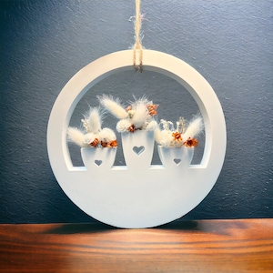 Könnte beinhalten: Weißer Holz-Kreis mit drei kleinen weißen Töpfen mit getrockneten Blumen, die an einem Seil hängen. Die Töpfe haben herzförmige Ausschnitte.