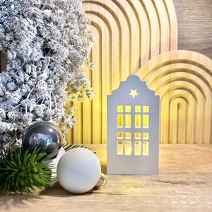Könnte beinhalten: Weißes, hausförmiges Licht aus Holz mit einem Stern oben und einem warmen gelben Schein aus den Fenstern. Das Licht steht auf einer Holzoberfläche mit einem weißen und grauen Weihnachtsbaumschmuck und einem grünen Tannenzweig.