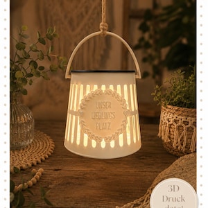 Puede incluir: Una lámpara decorativa blanca con forma de farolillo, con asa y cuerda para colgar. La lámpara tiene cortes verticales y un emblema decorativo con el texto "UNSER LIEBLINGS PLATZ". La luz está encendida, emitiendo un brillo cálido.