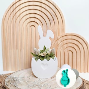 Pode incluir: Vaso de cerâmica branco em forma de ovo com um coração recortado, com a forma de um coelho. O vaso está cheio de flores secas e está sobre uma fatia de madeira. No canto inferior direito da imagem, é mostrado um cortador de biscoitos de coelho de plástico verde.