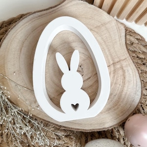 Könnte beinhalten: Weißer hölzerner Osterei-Ausschnitt mit einer Hasensilhouette im Inneren. Der Hase hat einen kleinen Herz-Ausschnitt in seiner Brust.