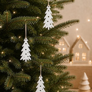 Könnte beinhalten: Drei weiße, flache, baumförmige Anhänger hängen an einem grünen Tannenbaum. Jeder Anhänger weist eine Reihe kreisförmiger Ausstanzungen entlang der Mitte und der Ränder auf und ist an einer dünnen, hellen Schnur zum Aufhängen befestigt.
